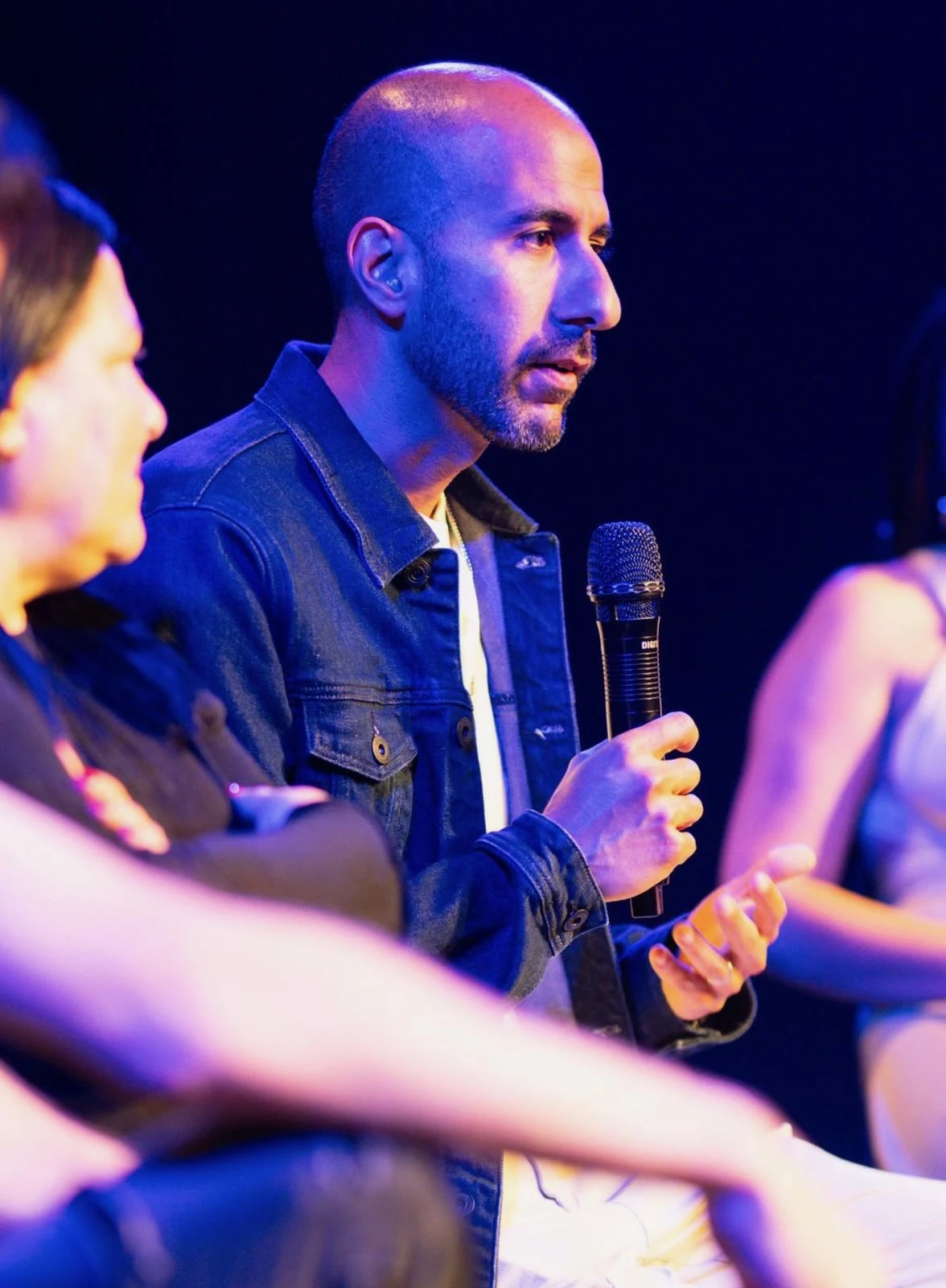 Haviv Ohayon speaking into a microphone during a blue-lit panel discussion.